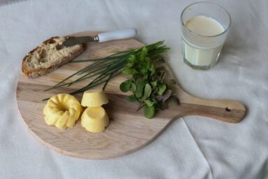 Hier zie je 3 vormpjes zelfgemaakte roomboter op een snijplank met een besmeerde snee brood en verschillende kruiden