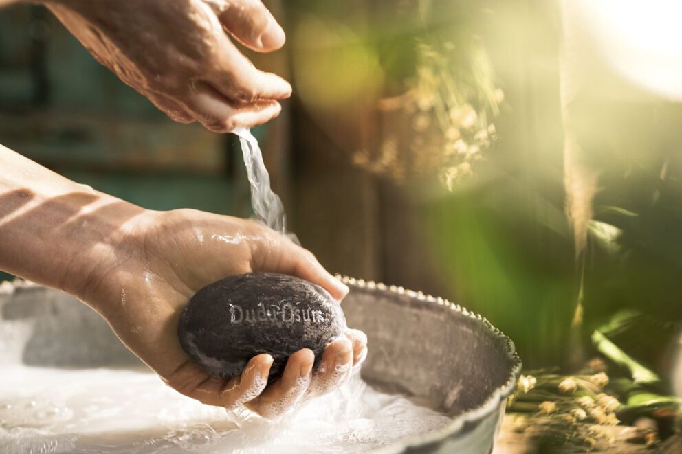 Een stuk zwarte zeep ligt in de hand boven een teiltje met water